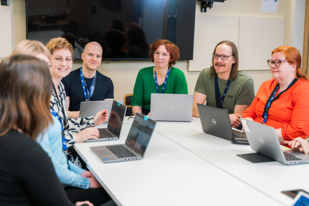 Seitsemän henkilöä kokoontuneena pöydän äärellä kannettavien tietokoneiden kanssa.