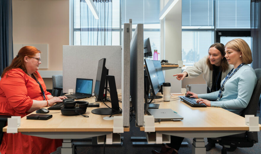 Avokonttorissa kaksi työpöytää peräkkäin, kolme henkilöä työskentelemässä tietokoneella.