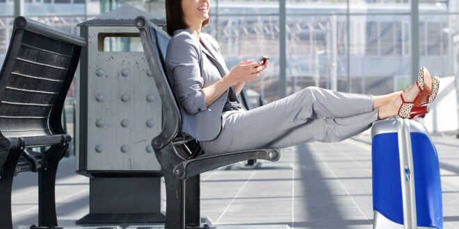 Henkilö istuu tuolilla odotusaulassa jalat matkalaukun päällä.
