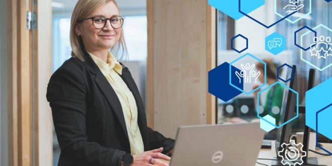 Hymyilevä nainen seisoo työpöydän ääressä. Hänen edessään on kannettava tietokone.