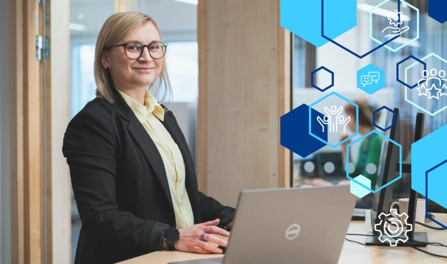 Hymyilevä nainen seisoo työpöydän ääressä. Hänen edessään on kannettava tietokone.